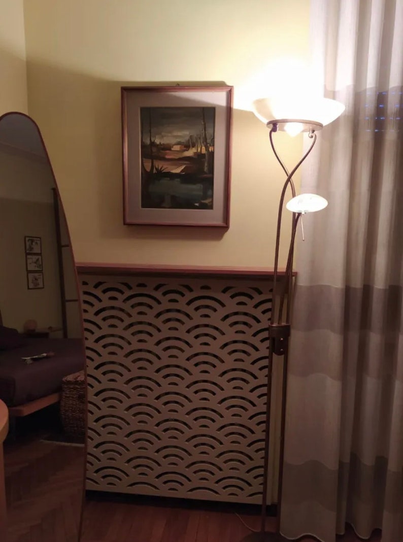 K&ouml;nnte beinhalten: Ein wei&szlig;er Heizk&ouml;rperverkleidung mit einem dekorativen Wellenmuster. Die Verkleidung ist an einer Wand mit einem gerahmten Gem&auml;lde dar&uuml;ber montiert. Eine Stehlampe mit einem wei&szlig;en Schirm steht neben der Verkleidung.