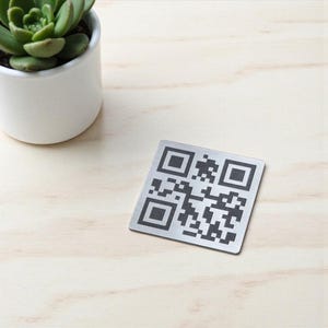 Può includere: Un'etichetta con codice QR quadrata color argento con un motivo nero e grigio. L'etichetta è su una superficie di legno chiaro, con un piccolo vaso di ceramica bianca che contiene una pianta succulenta verde nell'angolo in alto a sinistra.