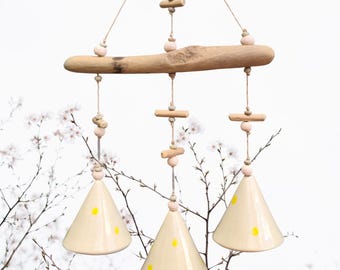Könnte beinhalten: Ein hängendes Windspiel mit einem Naturholzbalken und drei cremefarbenen Keramikglocken mit gelben Tupfen. Die Glocken sind mit Schnur aufgehängt, mit Holzakzenten und rosa Perlen. Dekorative Wohnkultur.