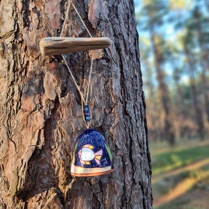 May include: A blue ceramic wind chime with a painted fish design hangs from a tree branch. The chime is suspended by a string with blue beads. The background is a blurred image of a forest.