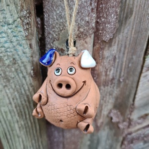 Può includere: Un ornamento di ceramica marrone a forma di maiale con dettagli dipinti in blu e bianco. Il maiale ha un viso sorridente ed è appeso a una corda.