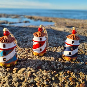 Op de afbeelding: Drie kleine keramische vuurtoren ornamenten met rode, blauwe en witte strepen. De ornamenten staan op een rotsachtig strand.