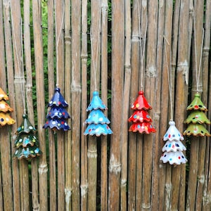 Könnte beinhalten: Eine Sammlung von Weihnachtsbaumschmuck aus Keramik in verschiedenen Farben, darunter Gelb, Grün, Blau, Rot und Weiß. Jeder Baum hat ein einzigartiges Design mit bunten Punkten und ist an einer Schnur aufgehängt. Die Ornamente werden vor einem Bambuszaun präsentiert.
