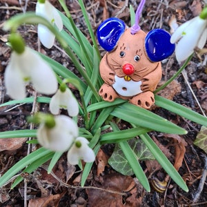 Peut inclure: Une figurine de souris en céramique brune avec des oreilles bleues et un nez rouge. La souris tient un cercle blanc dans ses pattes. La figurine est suspendue à une ficelle violette et est entourée de fleurs blanches de perce-neige.