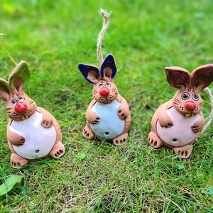 Può includere: Tre statuine di coniglio in ceramica con orecchie e pancia di colori diversi. I conigli sono marroni con nasi rossi e sono seduti su un letto di erba verde.