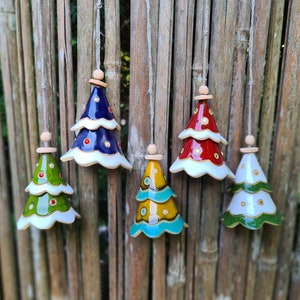 Op de afbeelding: Vijf keramische kerstboomornamenten in verschillende kleuren, waaronder groen, blauw, rood en geel. Elke boom heeft een witte rand en is opgehangen aan een touwtje. De ornamenten zijn versierd met stippen en hangen tegen een bamboe achtergrond.