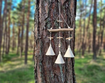 Könnte beinhalten: Ein handgefertigter Windspiel mit drei weißen, glockenförmigen Keramikanhängern, die an einem Treibholzstab hängen. Die Windspiele sind mit kleinen Holzperlen verziert und hängen vor einem verschwommenen Waldbild, was eine natürliche, rustikale Ästhetik erzeugt.