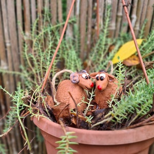 May include: Two small, brown, ceramic reindeer figurines are nestled in a hanging terracotta pot filled with green foliage. The reindeer have large, black eyes and red noses.