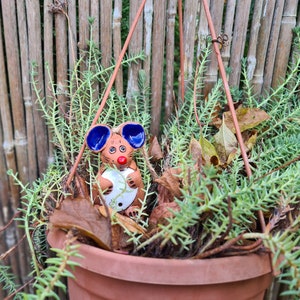 以下が含まれることがあります： 緑色の多肉植物が入ったテラコッタの吊り鉢と、陶器製のネズミの置物。 ネズミは青い耳と赤い鼻をしており、白いハート型のオブジェクトを持っています。 背景には竹のフェンスがあります。