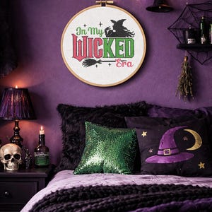 Pode incluir: Um bastidor de bordado redondo com o texto "In My Wicked Era" em vermelho e verde, com uma silhueta de bruxa e uma vassoura. Está pendurado numa parede roxa, decorada com chapéus de bruxa e luzes de corda, com tema de Halloween.