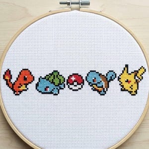 May include: Cross-stitch embroidery featuring five colorful Pokémon characters: Charmander, Bulbasaur, a Poké Ball, Squirtle, and Pikachu. The design is set against a white fabric background, framed by a wooden embroidery hoop.