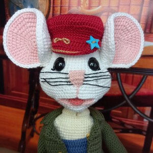 May include: A white crocheted mouse wearing a red crocheted hat with a gold trim and a blue star. The mouse has black whiskers and pink ears.