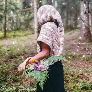 Könnte beinhalten: Eine Person trägt einen gestreiften, gehäkelten Schal mit Kapuze in einer Waldlandschaft. Der Schal ist in Braun- und Weißtönen gehalten. Die Person hält einen Korb mit Blumen und Farnen.
