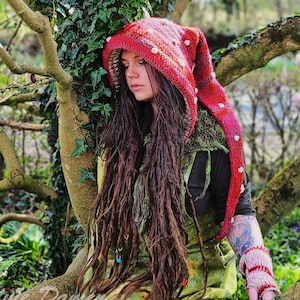 Może przedstawiać: Kobieta z długimi brązowymi dredami nosi czerwony i biały kaptur z białymi kropkami. Kaptur ma długi, spiczasty czubek. Siedzi na drzewie z zielonymi liśćmi.