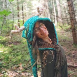 Puede incluir: Un gorro de punto de color azul turquesa con cuernos marrones y beige. El gorro tiene una cola larga y lo lleva puesto una persona en un entorno boscoso.