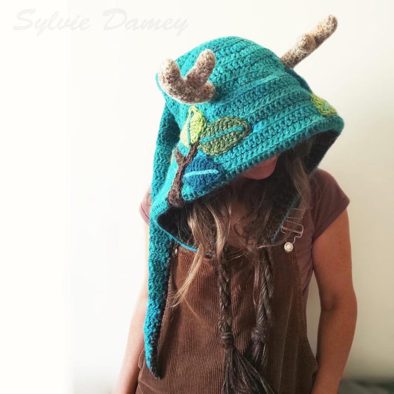Puede incluir: Un gorro de punto de color azul turquesa con capucha y cola larga, adornado con cuernos marrones y hojas verdes. El gorro lo lleva puesto una persona con el pelo largo y casta&ntilde;o.
