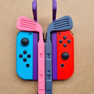 May include: Two Nintendo Switch controllers with custom 3D printed attachments. The controllers are connected by a purple and grey cable. The attachments are shaped like golf clubs and are pink and grey.
