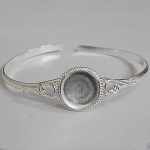 May include: Silver bangle bracelet with a round, bezel-set, blank cameo. The cameo is surrounded by a decorative border.