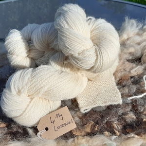 May include: A close-up of a skein of white 4-ply Lambswool yarn. The yarn is on a brown and white surface with a small piece of knitted fabric.