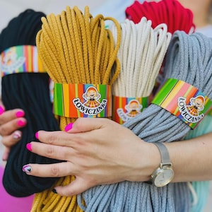 May include: Bundles of colorful macrame cord in black, mustard yellow, cream, gray, and red. Each bundle is wrapped with a colorful label featuring a smiling cartoon figure and the word "ŁOWICZANKA". The cords are held in a person's hands.
