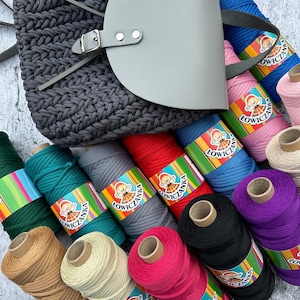Peut inclure: Un sac fourre-tout gris tissé avec un rabat et une sangle en cuir gris. Des bobines de fil coloré de différentes nuances, dont le vert, le rouge, le bleu, le rose et le violet, sont disposées autour du sac. Chaque bobine a une étiquette colorée.