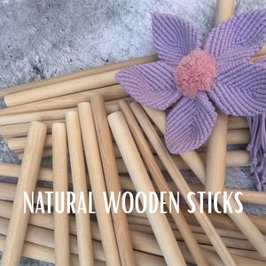Op de afbeelding: Een verzameling natuurlijke houten stokken, variërend in lengte. Een grote, lavendelkleurige macramé bloem met een roze centrum is bovenop enkele van de stokken geplaatst. De tekst "NATURAL WOODEN STICKS" wordt in het wit weergegeven.
