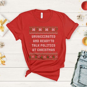 以下が含まれることがあります： 白と緑のクリスマスセーターのデザインが施された赤いTシャツ。シャツのテキストは「ワクチン未接種でクリスマスに政治について話す準備万端」です。
