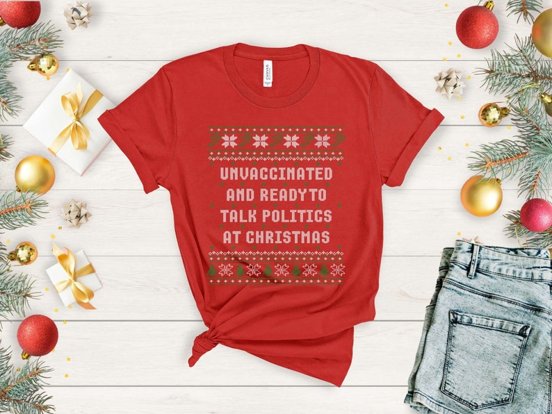 以下が含まれることがあります： 白と緑のクリスマスセーターのデザインが施された赤いTシャツ。シャツのテキストは「ワクチン未接種でクリスマスに政治について話す準備万端」です。