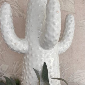 May include: A white ceramic cactus ornament with a textured surface and multiple arms. The cactus is a decorative item, and is paired with green leaves and brown olives. The background features a floral pattern.