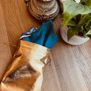 May include: A gold drawstring bag with a teal blue and white patterned fabric inside. The bag is sitting on a wooden surface.