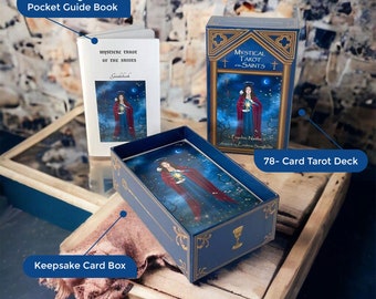 May include: A blue and gold box with a 78-card tarot deck inside. The box has a gold trim and the words "Mystical Tarot of the Saints" on the front. The deck is illustrated with a woman holding a lantern. The box is sitting on a wooden surface with a brown fabric next to it. A 96-page guidebook is also visible.