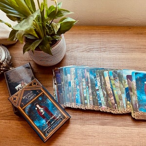 May include: A deck of Mystical Tarot Saints cards with a blue, gold, and white design. The cards are spread out on a wooden table. The box for the cards is open and shows a woman in a blue and red robe with a crown on her head. The text on the box reads "Mystical Tarot Saints: Angels & Enchanting Spirits".