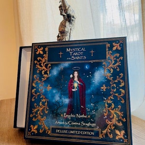 May include: A blue box with gold accents and the text "Mystical Tarot of the Saints" and "Psychic Nathe" with artwork by "Cristina Staglioti" and "Deluxe Limited Edition".