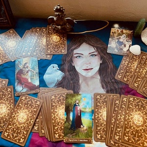 May include: A tarot card deck with gold accents and intricate designs. The cards are spread out on a blue fabric with a white dove and a figure of a woman. The cards feature a variety of images, including a figure in a red robe, a figure in a blue robe, and a figure with a staff.