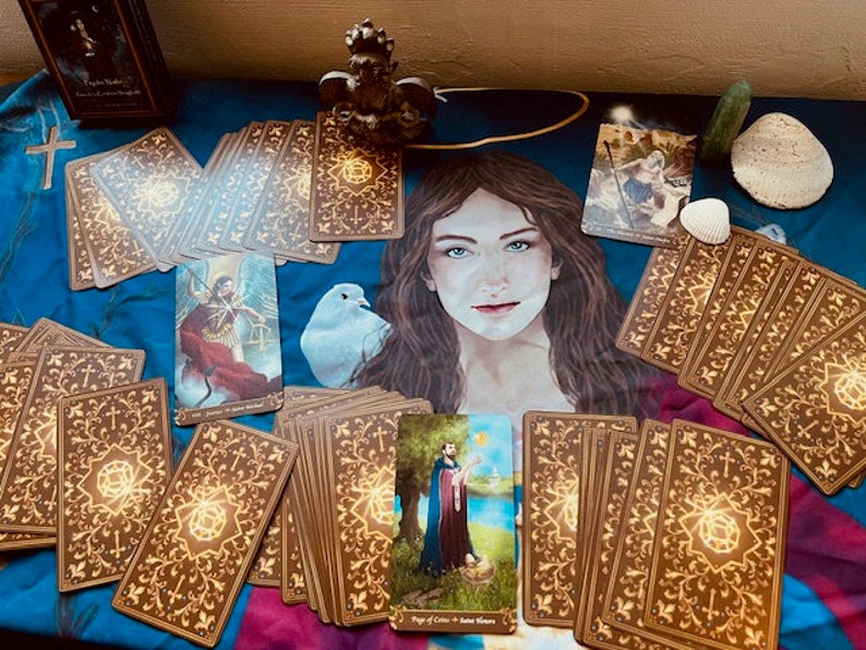 May include: A tarot card deck with gold accents and intricate designs. The cards are spread out on a blue fabric with a white dove and a figure of a woman. The cards feature a variety of images, including a figure in a red robe, a figure in a blue robe, and a figure with a staff.