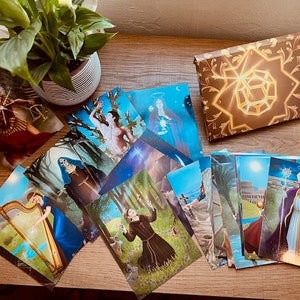 May include: A set of tarot cards with colorful illustrations and gold accents. The cards are laid out on a wooden table with a plant in a pot in the background. The box for the cards is closed and features a gold design with a geometric shape in the center.
