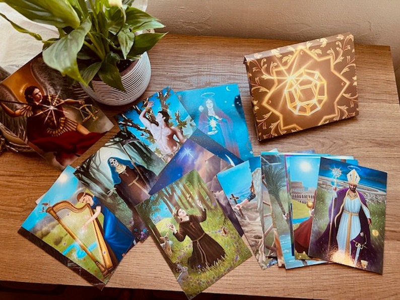 May include: A set of tarot cards with colorful illustrations and gold accents. The cards are laid out on a wooden table with a plant in a pot in the background. The box for the cards is closed and features a gold design with a geometric shape in the center.