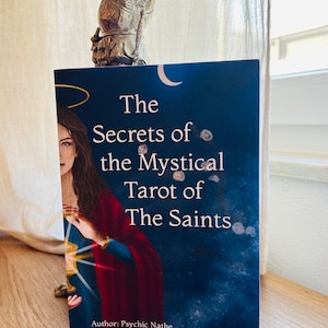 May include: A book titled "The Secrets of the Mystical Tarot of The Saints" with a blue cover and a painting of a woman in a red and blue robe holding a golden chalice. The author is Psychic Nathe and the illustrations are by Cristina Scagliotti.