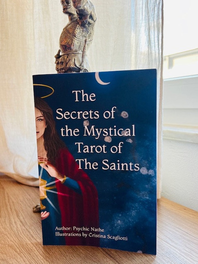 May include: A book titled "The Secrets of the Mystical Tarot of The Saints" with a blue cover and a painting of a woman in a red and blue robe holding a golden chalice. The author is Psychic Nathe and the illustrations are by Cristina Scagliotti.
