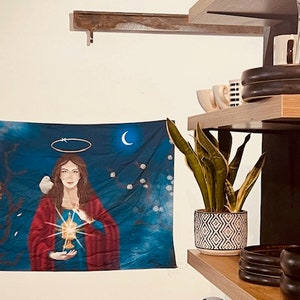 May include: A blue tapestry with a painting of a woman holding a chalice with a glowing light. The woman is wearing a red robe and has a halo above her head. A white dove is perched on her shoulder. The tapestry is hanging on a white wall.