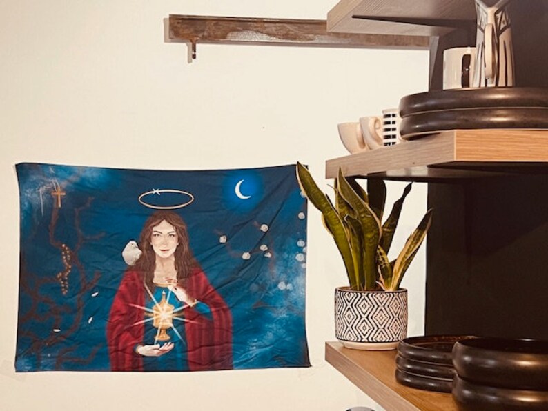 May include: A blue tapestry with a painting of a woman holding a chalice with a glowing light. The woman is wearing a red robe and has a halo above her head. A white dove is perched on her shoulder. The tapestry is hanging on a white wall.