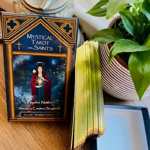 May include: A deck of Mystical Tarot of the Saints cards with gold gilded edges. The box is black with a gold trim. The deck features artwork by Cristina Scaglioti.