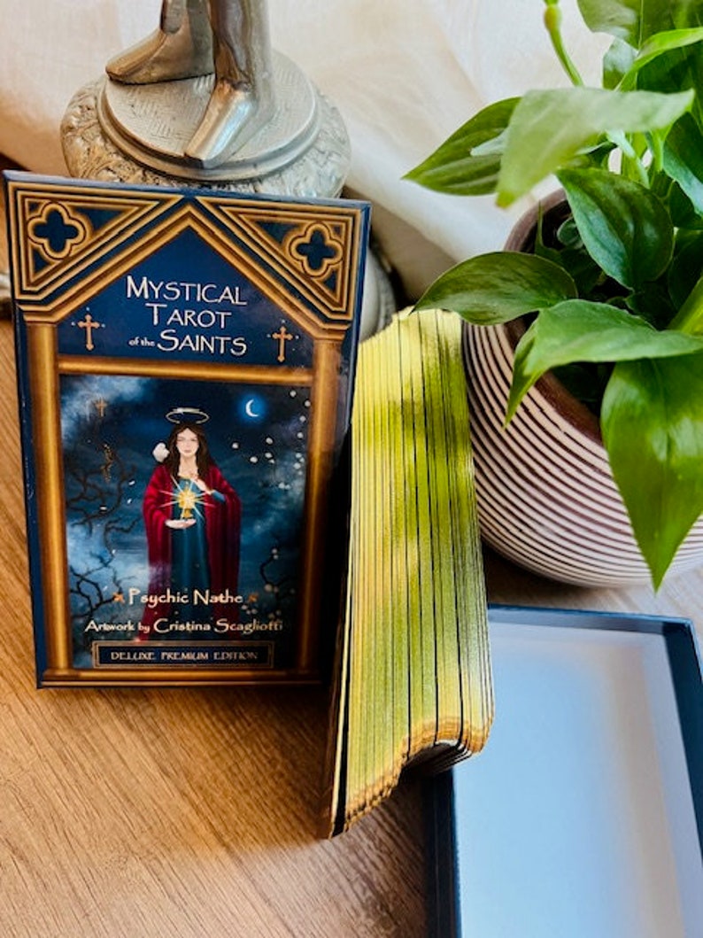 May include: A deck of Mystical Tarot of the Saints cards with gold gilded edges. The box is black with a gold trim. The deck features artwork by Cristina Scaglioti.