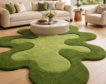 May include: A modern living room featuring a large, abstract-shaped green rug with varying shades of green. The room includes a beige sofa with green and beige pillows, a round coffee table, and a white armchair. Large windows offer a view of lush greenery.