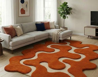 May include: An orange and cream-colored area rug with an abstract, flowing design. The rug is placed in a living room with a gray sectional sofa, throw pillows, and a potted plant. The rug's unique shape adds a modern touch to the room's decor.