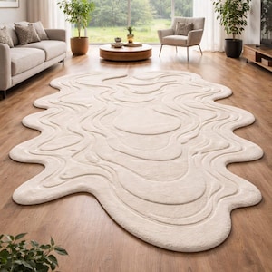 May include: A cream-coloured, abstract-shaped rug with a wavy design. The rug is placed on a hardwood floor in a living room setting. The room features a sofa, armchair, and plants, creating a cosy atmosphere.