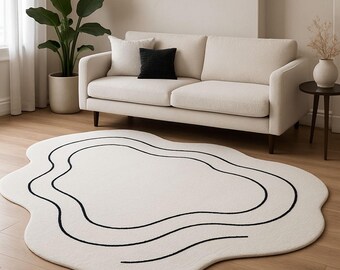 May include: A cream-colored, abstract-shaped rug with black wavy lines. The rug is placed on a hardwood floor in front of a cream-colored sofa. A black pillow and a white pillow are on the sofa.