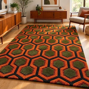 May include: A rectangular area rug featuring a repeating geometric pattern of interlocking hexagons in vibrant orange, deep olive green, and bold black. The rug has a thick, textured pile that creates a distinct visual depth across the floor surface.