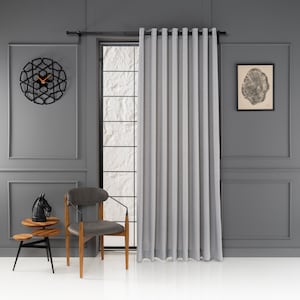 May include: A grey linen curtain with a black curtain rod hangs in front of a window. A black and white clock is on the wall to the left of the window. A wooden chair with a grey cushion sits in front of the window.