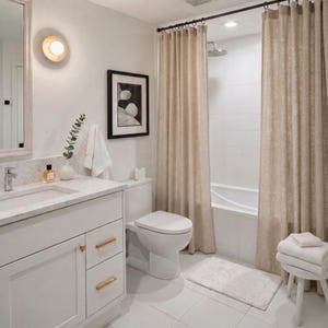 May include: A modern bathroom with white tile flooring and walls. Features include a white vanity with gold hardware, a toilet, and a shower area with beige curtains. A framed black and white art piece hangs on the wall, and a small white rug is on the floor.